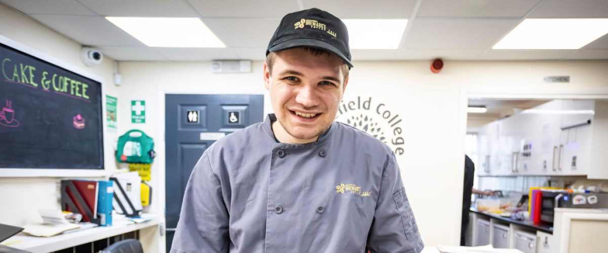 Male student in Breward's barista uniform smiling at camera while holding two plates of baked goods